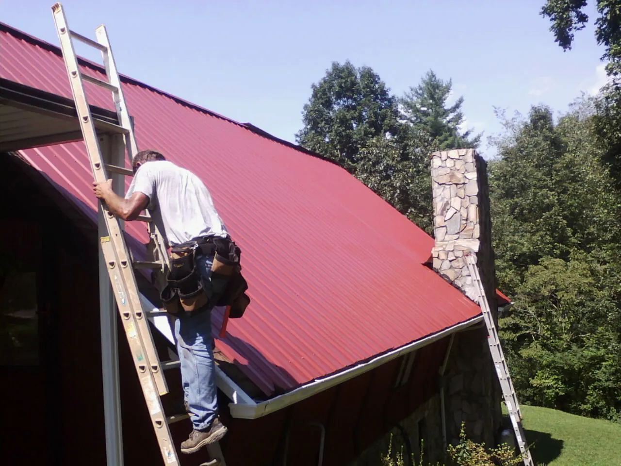 Roofer on a ladder beside a red metal roof and stone chimney — Soffit & Fascia Repair by Roofing Brothers in Inwood